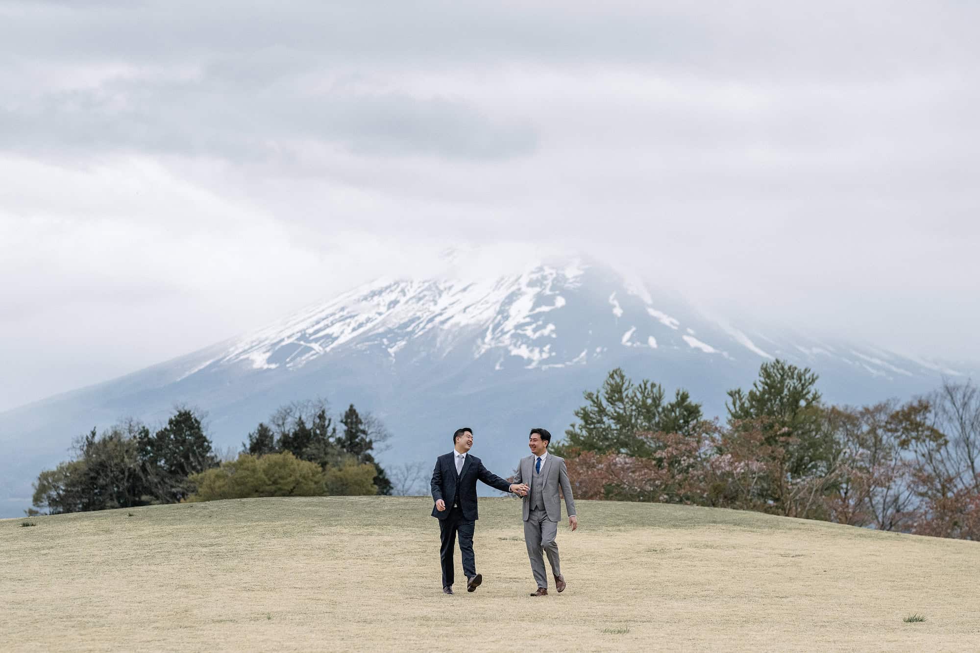 Mt Fuji engagement photos near Kawaguchiko, the couple laughing hand in hand across a grassy rise as the snowy summit hides in cloud behind.