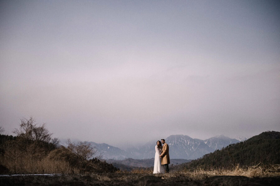 The bride and groom stand small on a dry winter hilltop under a pale sky on their Nagano winter wedding day, the snowy Northern Alps in cloud behind.