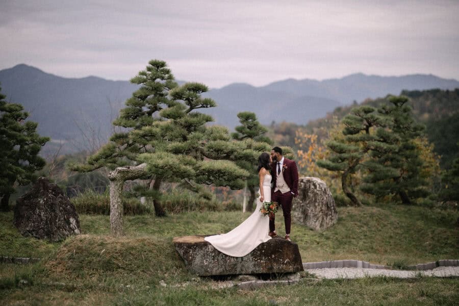 The bride and groom kiss on a flat rock among sculpted pines, her train across the stone, the Uda ranges layered behind their Nara wedding.