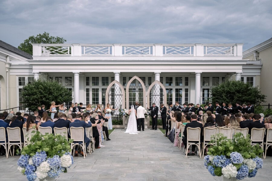 Guests stretch their hands toward the couple in prayer under a grey storm sky at their Piedmont Driving Club wedding, three Gothic arches behind.