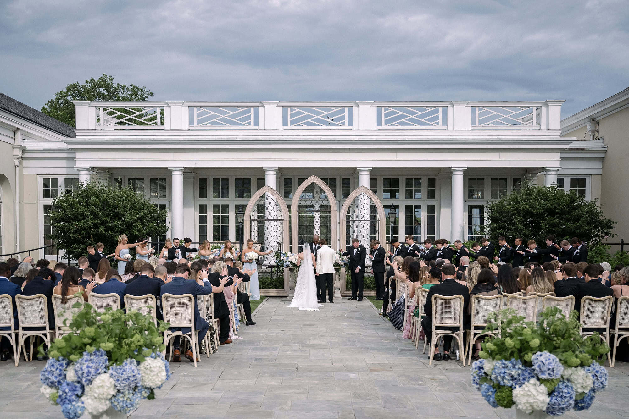 Guests stretch their hands toward the couple in prayer under a grey storm sky at their Piedmont Driving Club wedding, three Gothic arches behind.