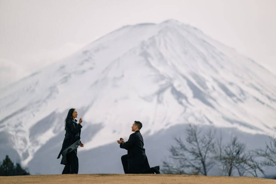 He kneels with the ring box as she laughs on a Kawaguchiko hilltop, the snow-covered mountain behind their surprise proposal at Mt Fuji.