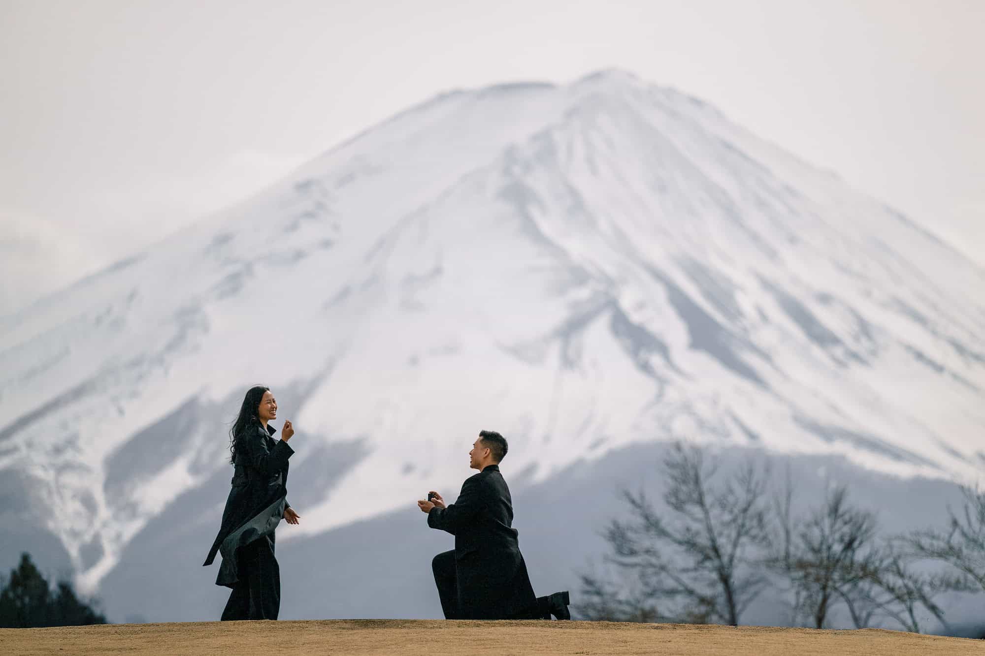 He kneels with the ring box as she laughs on a Kawaguchiko hilltop, the snow-covered mountain behind their surprise proposal at Mt Fuji.