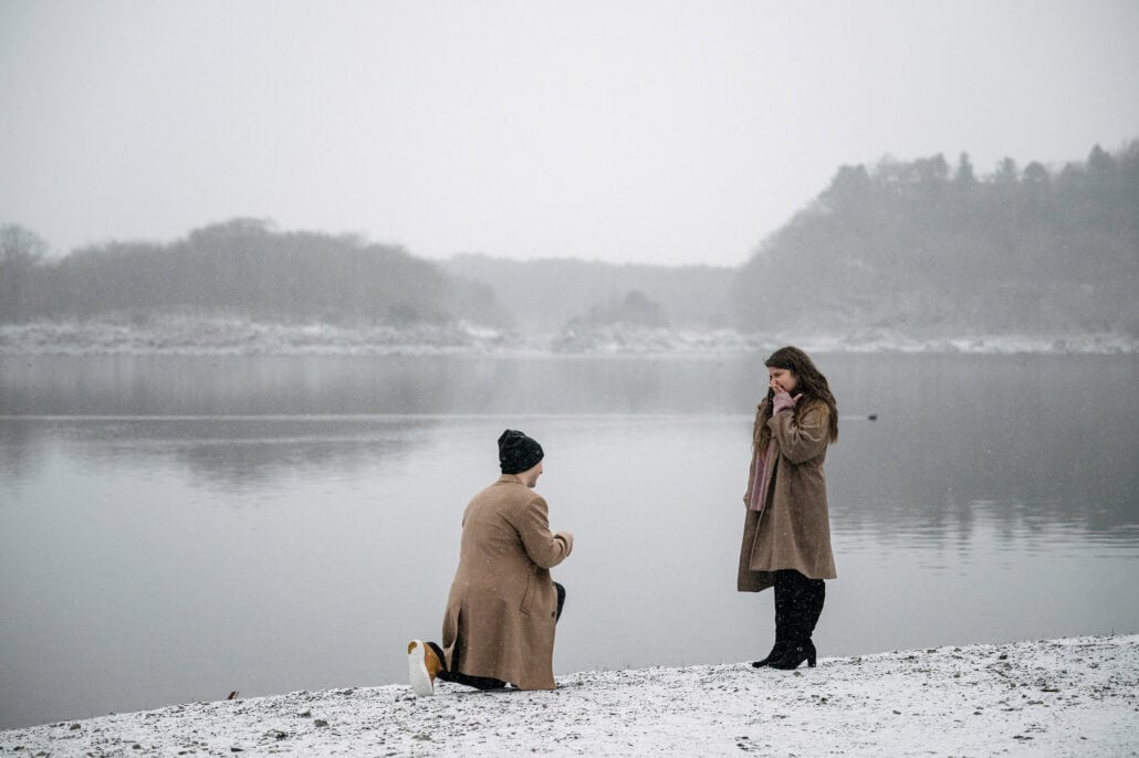 He kneels on one knee on the snowy shore as she lifts her hands to her face, a surprise proposal with Lake Shoji and its misty hills behind them.