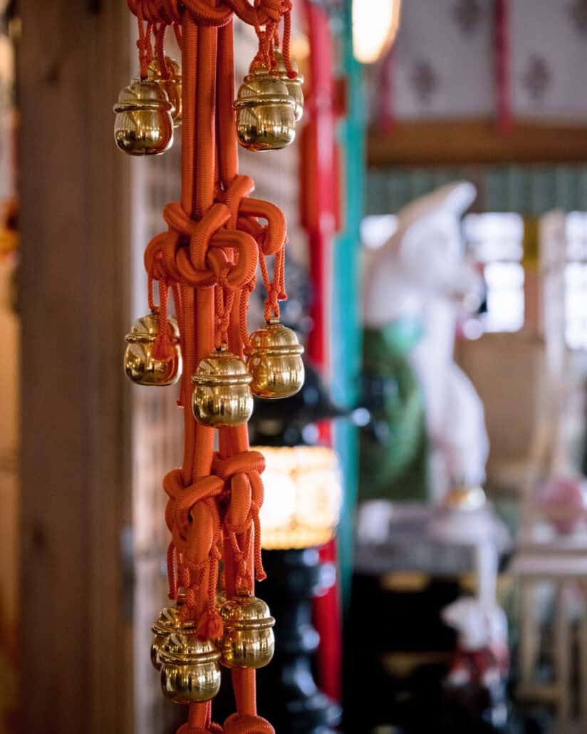 Golden bells hang from a red cord in Takayama Inari Shrine, a white fox statue blurred behind them, as the Shinto blessing begins.