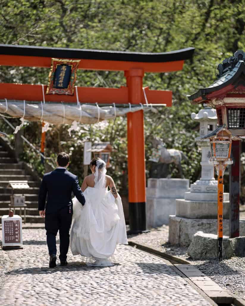 The couple walk toward the stone steps of Takayama Inari Shrine through its vermilion torii, stone lanterns lining the path.