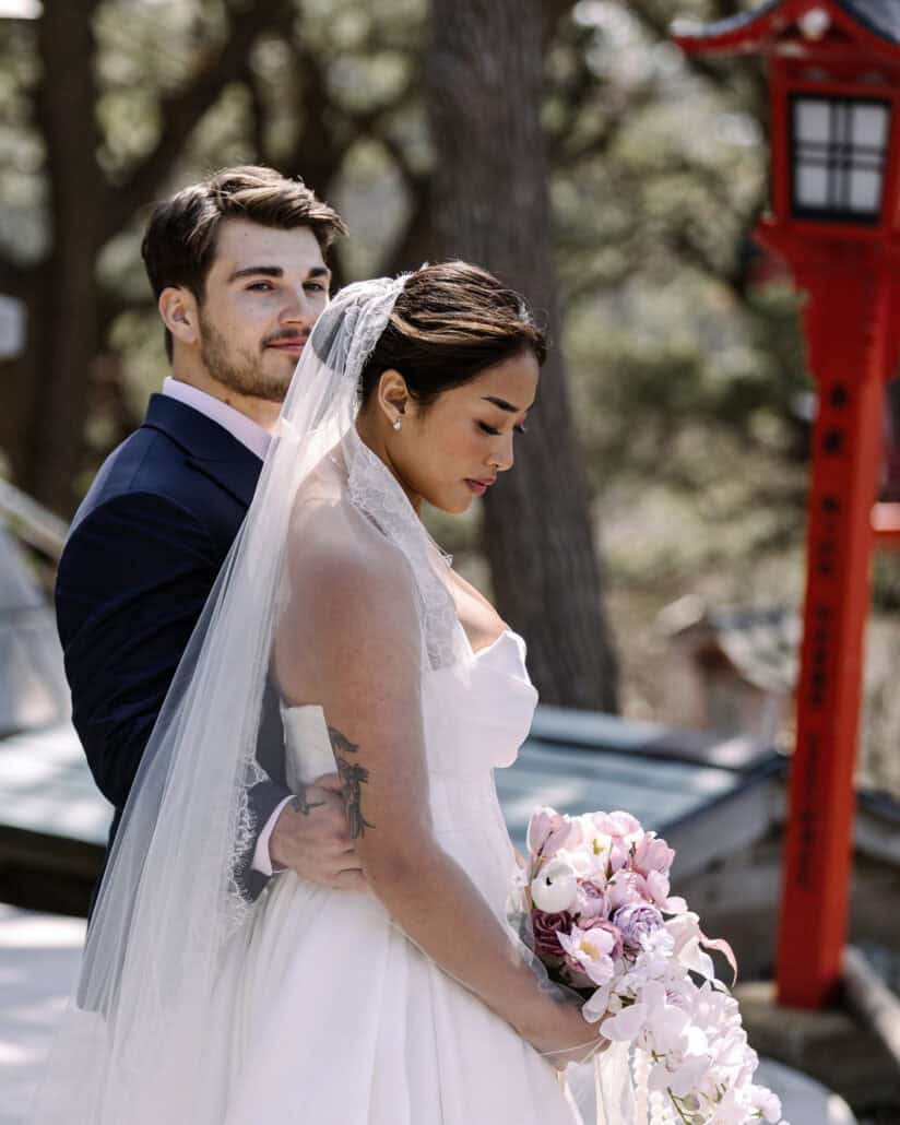 The groom holds the bride from behind beside a red lantern at Takayama Inari Shrine, her bouquet of pink orchids in her hands.