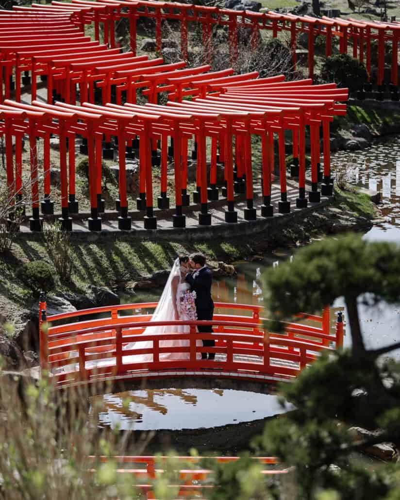The bride and groom embrace on the vermilion bridge as the rows of red torii curve along the pond at Takayama Inari Shrine.