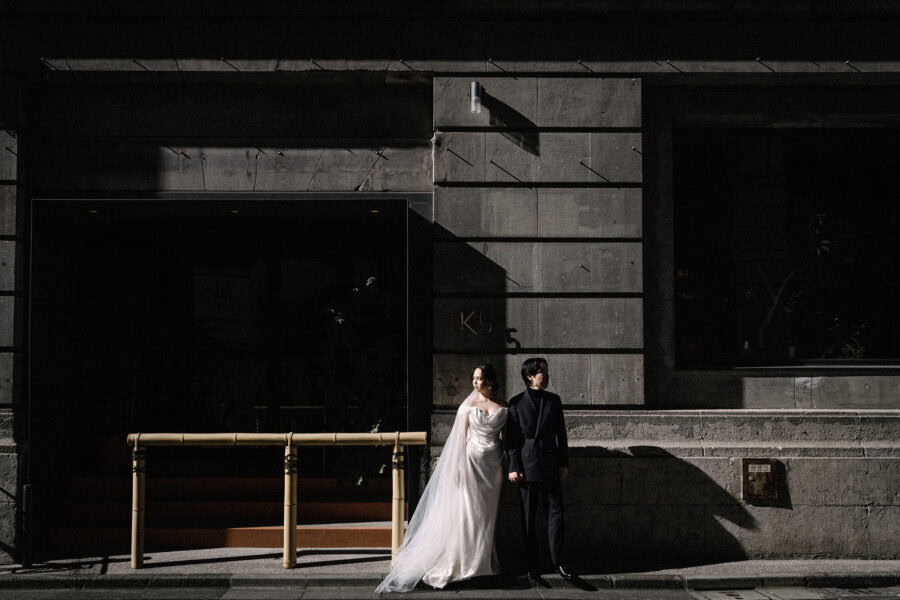 The bride and groom stand in a slice of hard sun against the dark stone of a 1923 bank building in Kabutocho on the day of their Tokyo Bay wedding.