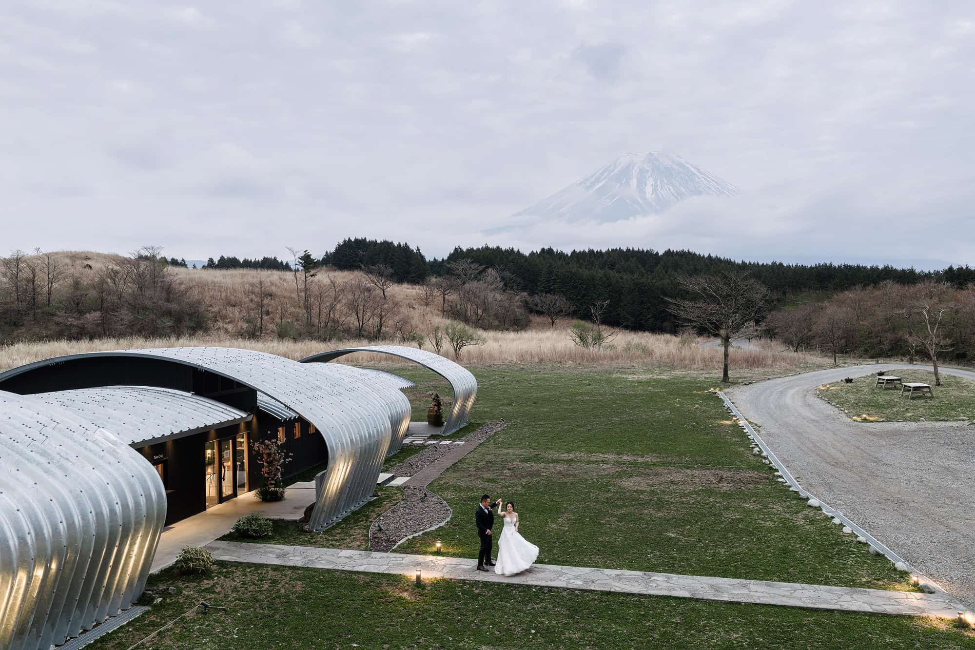 The groom twirls the bride on a lit path beside the silver vaults of Fujisan Winery, a unique wedding venue in Japan, as Mt Fuji clears the cloud.