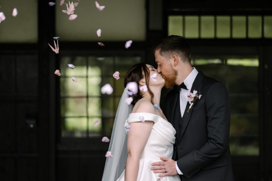 Paper cranes and pink petals fall around the bride and groom as they kiss in front of the dark timber hall of Genji-kan at Jinya.