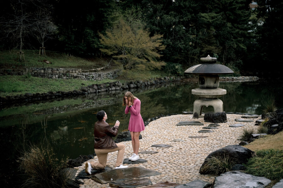 Beside a stone lantern at a quiet garden pond in Shinjuku, Tokyo, a man kneels as a woman in pink lifts her hands to her face.