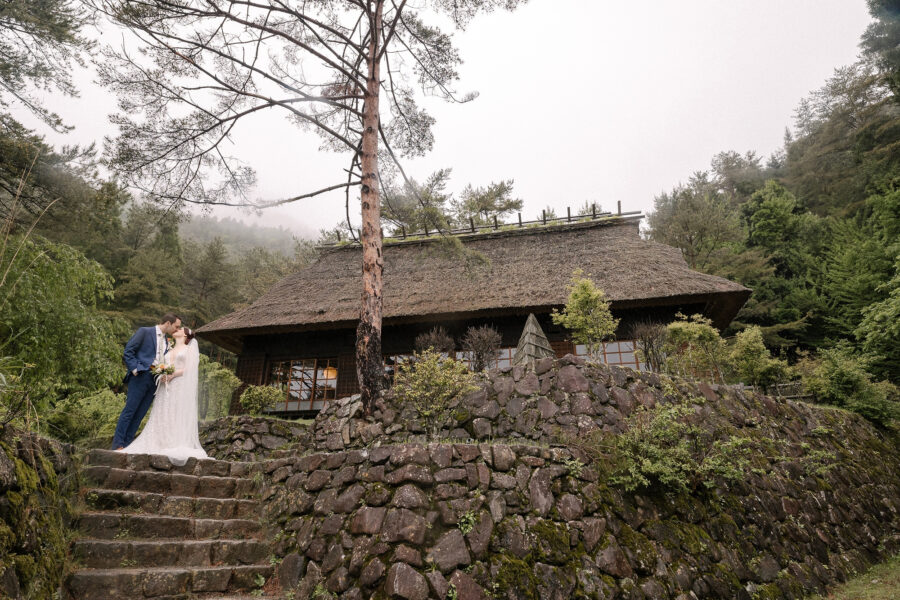 The couple kiss on mossy stone steps below a pine and a thatched farmhouse at Saiko Iyashi no Sato Nenba, with mist on the hills.