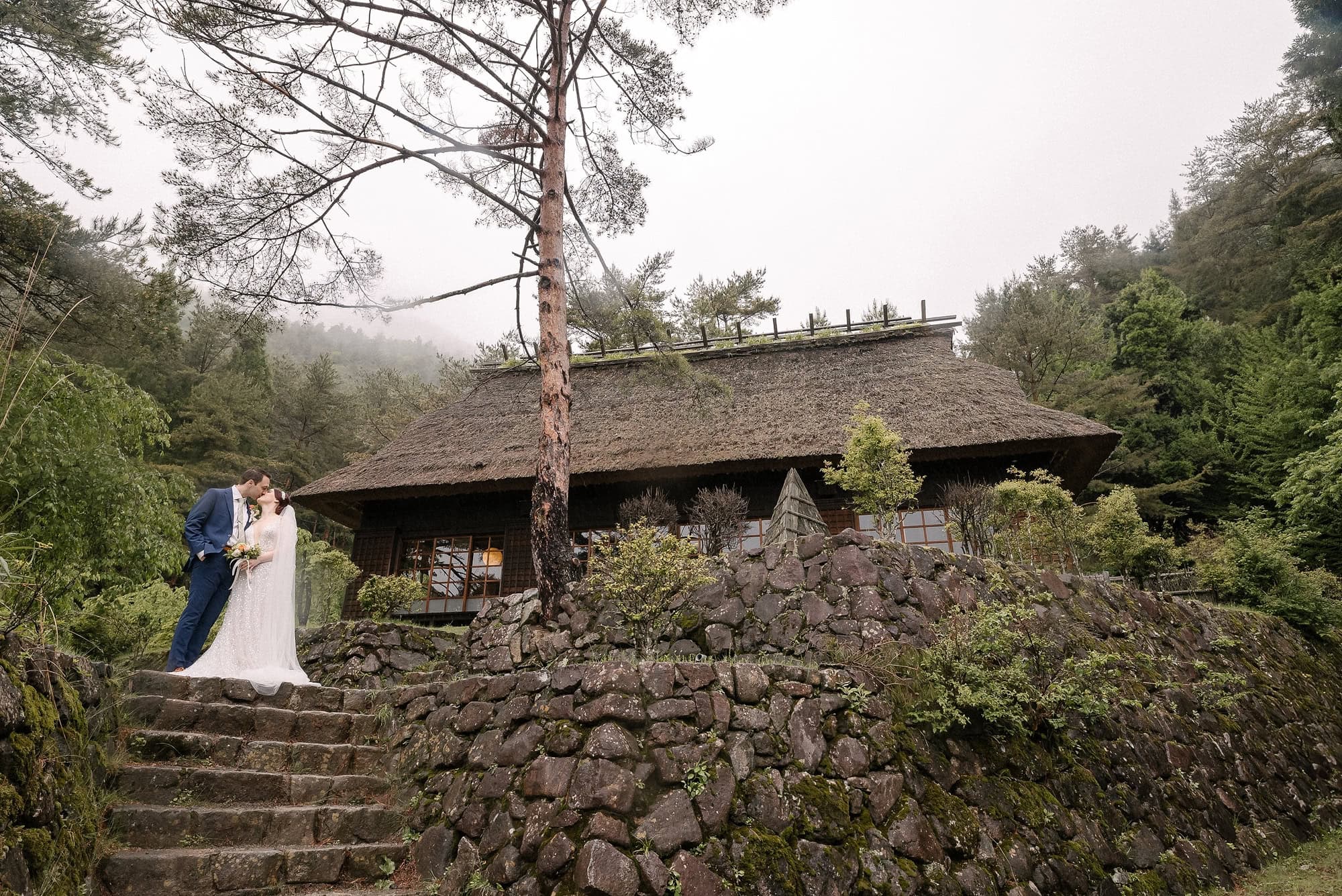 The couple kiss on mossy stone steps below a pine and a thatched farmhouse at Saiko Iyashi no Sato Nenba, with mist on the hills.