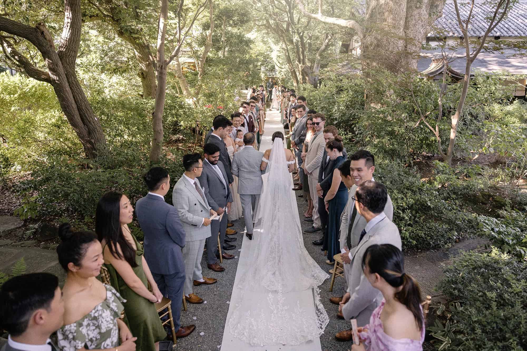 Guests line a white aisle under green trees on the forest walkway at Jinya for a destination wedding ceremony in Japan.