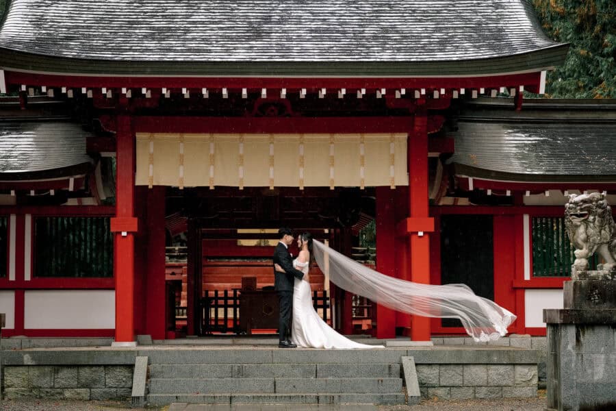 The bride's veil streams out in the wind as the couple stand before the vermilion gate of Fuji Omuro Sengen Jinja in autumn rain.