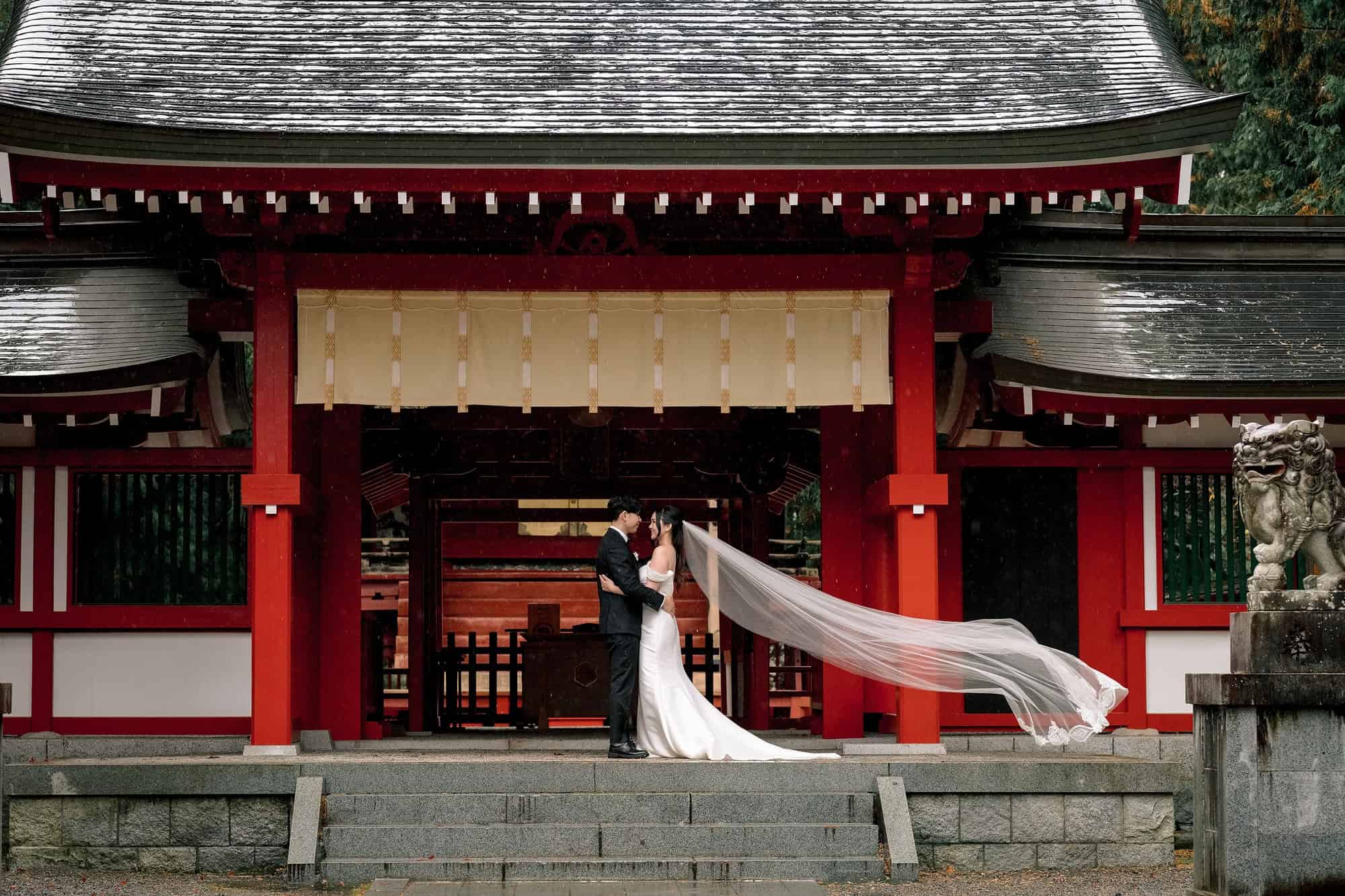 The bride's veil streams out in the wind as the couple stand before the vermilion gate of Fuji Omuro Sengen Jinja in autumn rain.