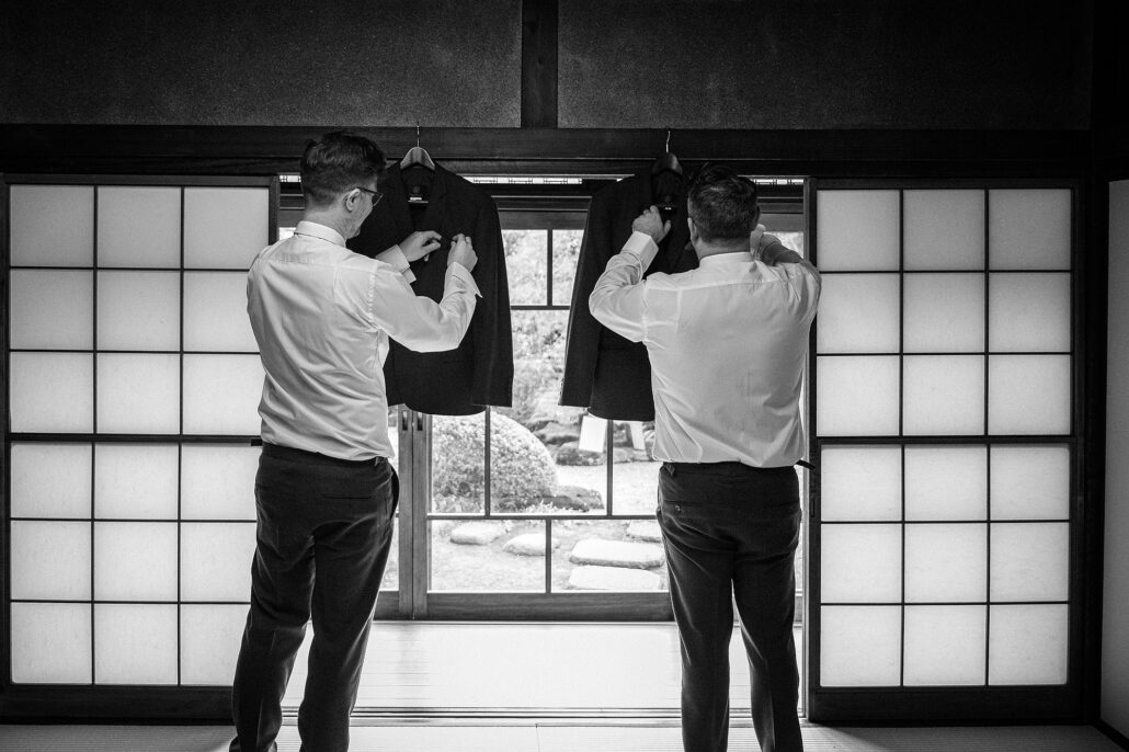 Two grooms hang their suit jackets in the shoji doorway of an old house at Tantoku in Kawagoe, the winter garden beyond the glass.