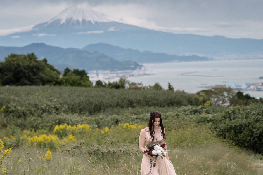 The bride stands with her bouquet in the Nihondaira tea fields, Mt Fuji and Suruga Bay behind her, at a Mt Fuji tea field elopement.