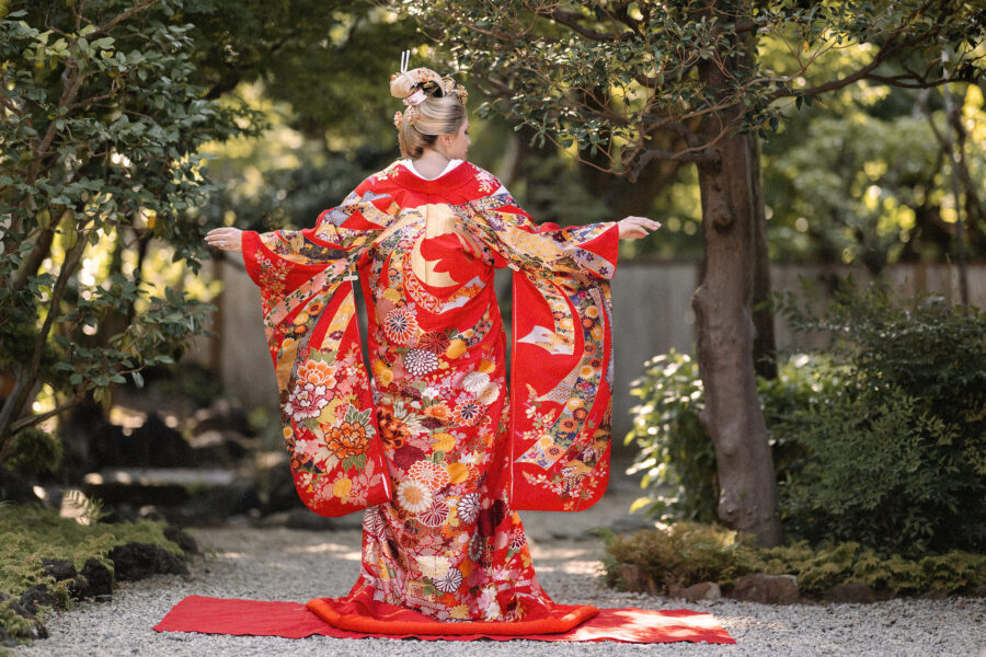 Bride in a red iro-uchikake wedding kimono and nihongami, arms open to show the back of the robe, on a gravel path in a Tokyo garden
