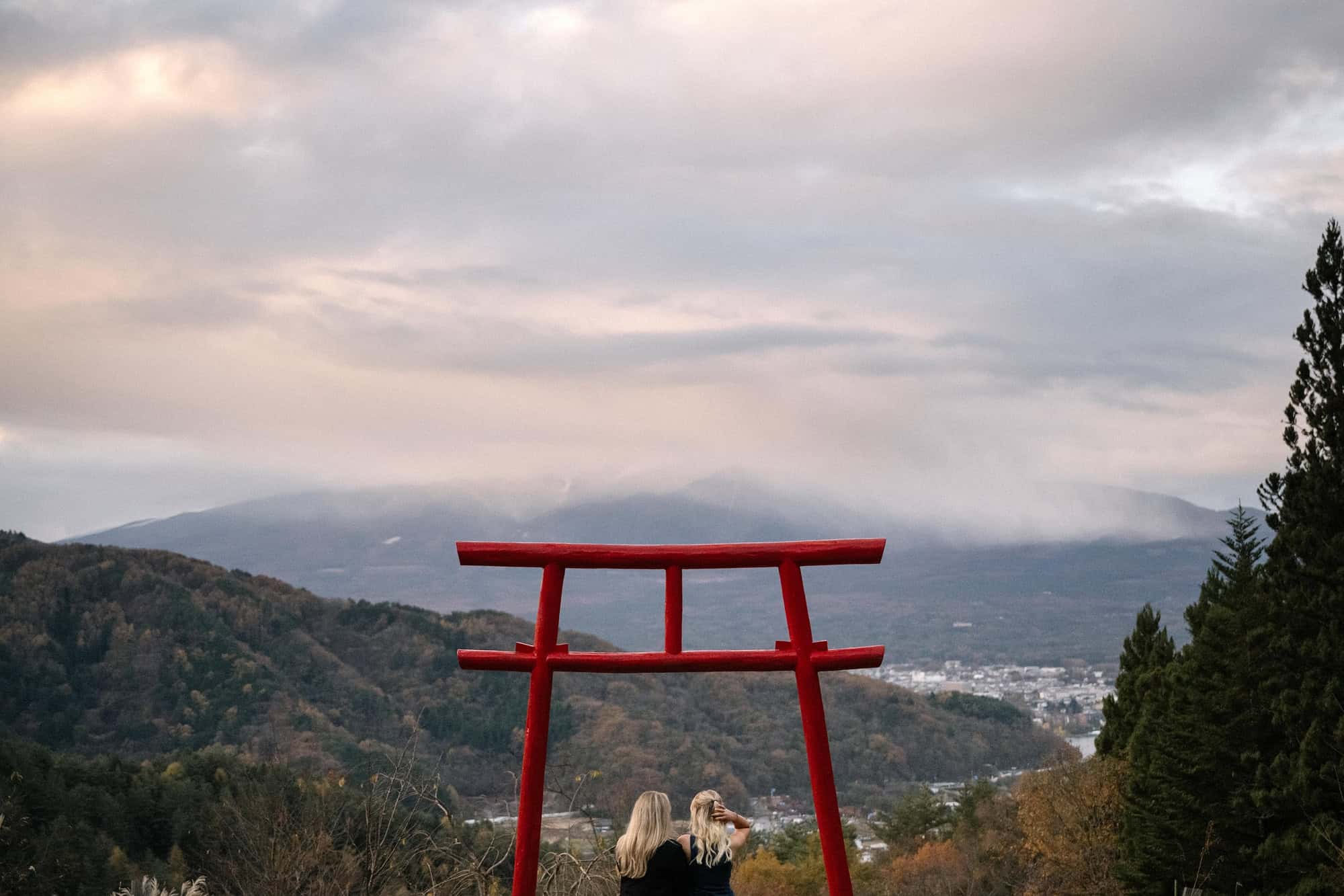 Two women stand small beneath a vermilion torii on a hillside above Kawaguchiko, a proposal in Japan photographed with the shrine's permission.