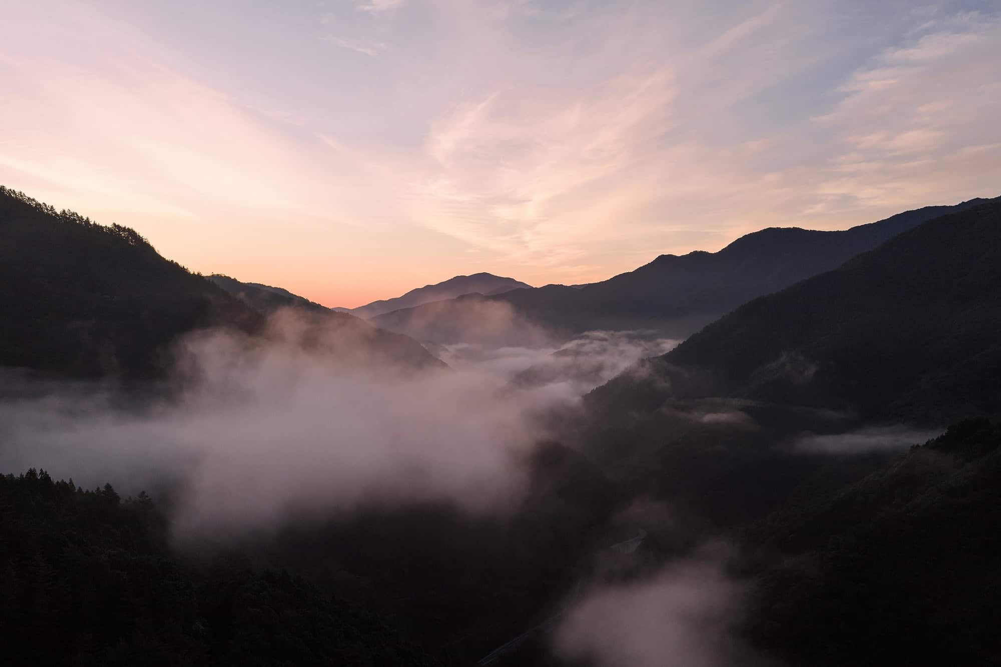 Cloud fills the forested valleys around a mountain village in Yamanashi at dawn, pink light on the ridges, the morning of a June elopement in Japan.