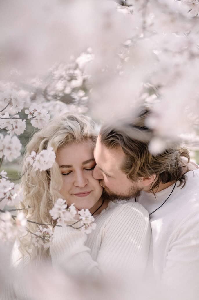 Eyes closed, the bride-to-be leans into her fiance's kiss on her cheek, white cherry blossom framing them in a Tokyo park.