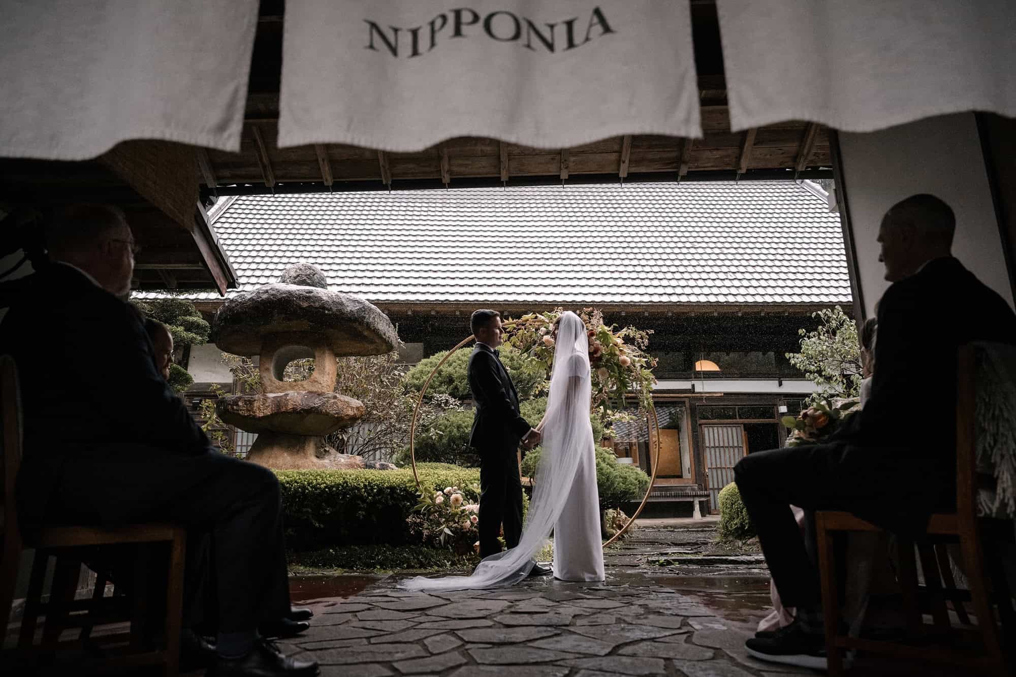 Guests sit under the eaves and noren of an old house in Kosuge as the couple marry in the garden beyond, a wedding venue in Japan.