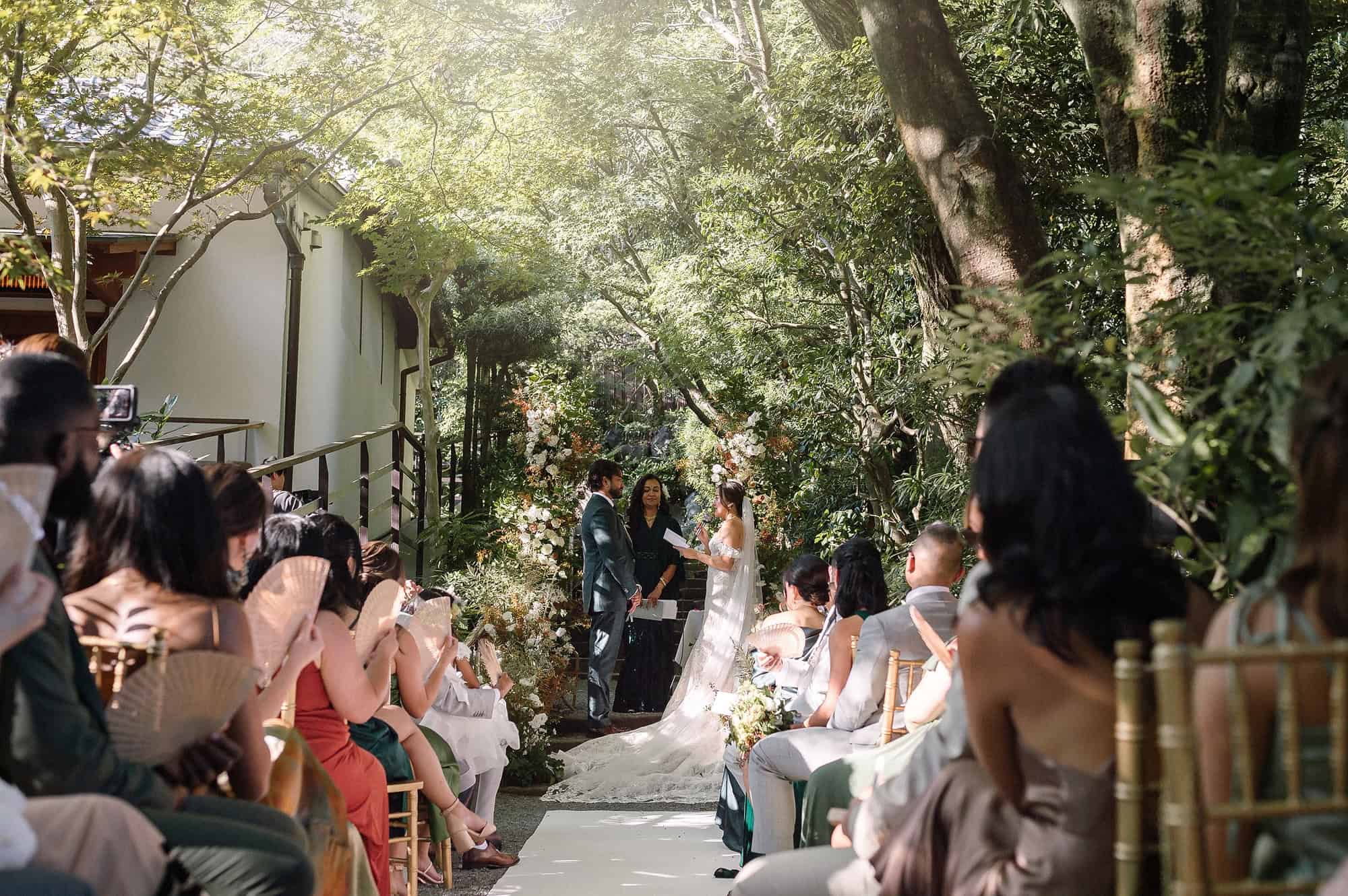 Guests sit on gold chairs under the trees either side of the walkway aisle at Jinya, an exclusive use wedding venue in Japan.