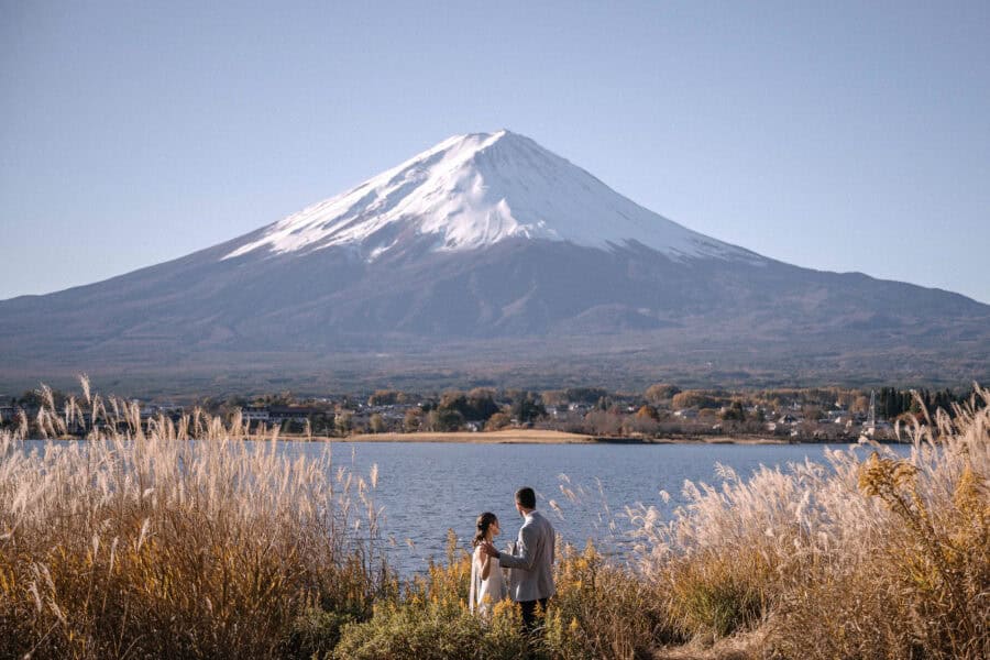 The bride and groom stand in silver grass on the shore of Lake Kawaguchiko, with the whole of Mount Fuji clear across the water.