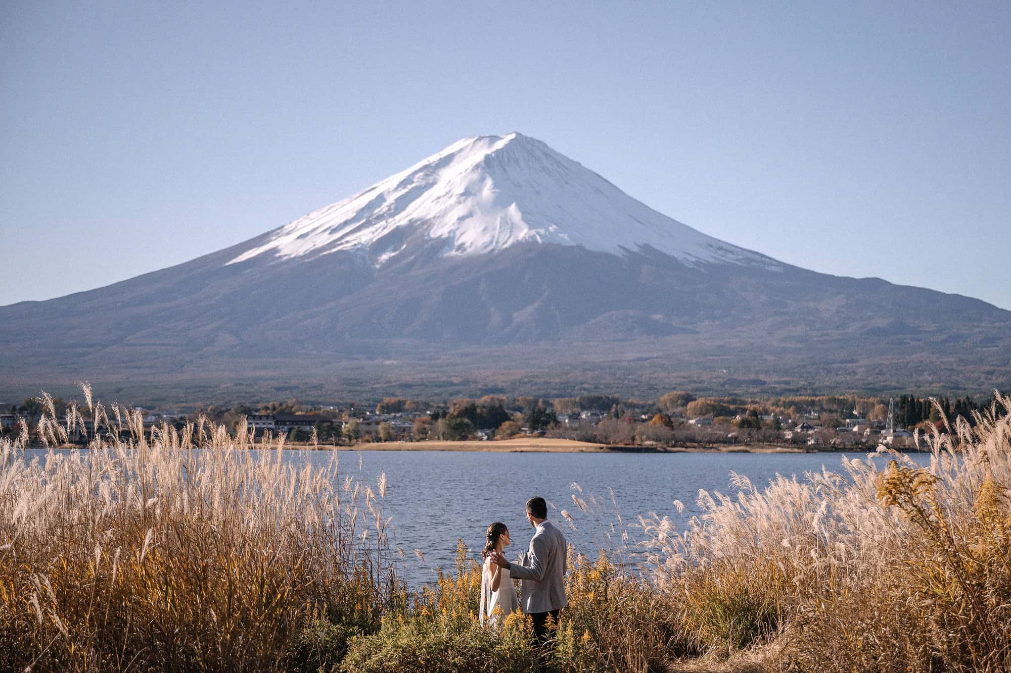 The bride and groom stand in silver grass on the shore of Lake Kawaguchiko, with the whole of Mount Fuji clear across the water.