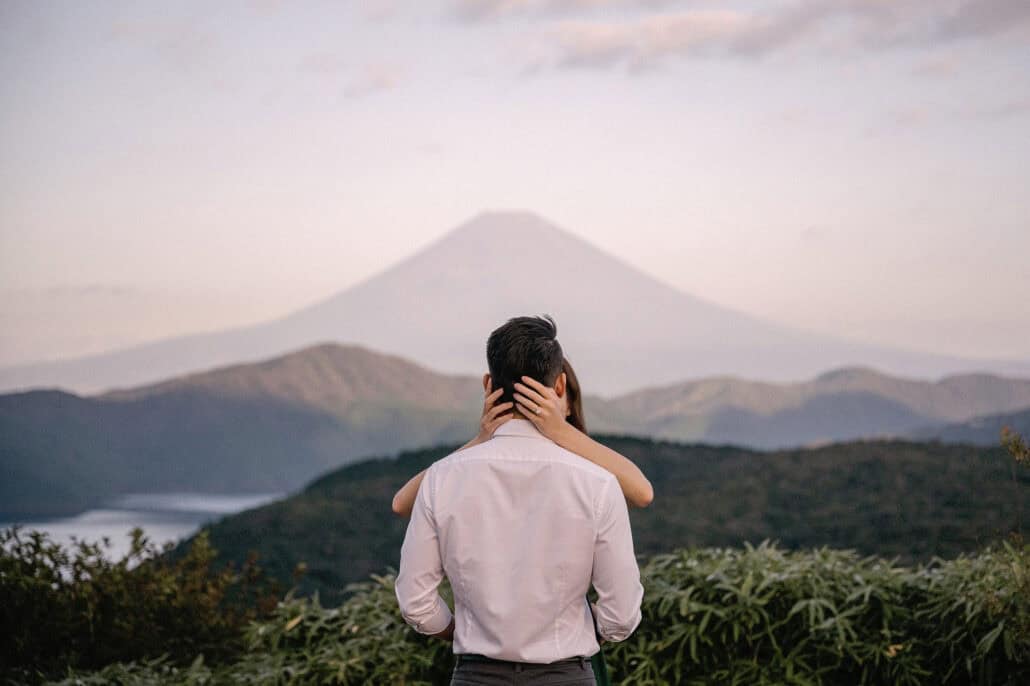 She reaches around him from behind as they face Mt Fuji across the hills above Lake Ashi in soft pink morning light.