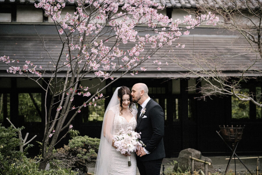 The groom kisses the bride's forehead under a pink cherry tree beside a dark timber hall, at their intimate spring wedding in Japan.