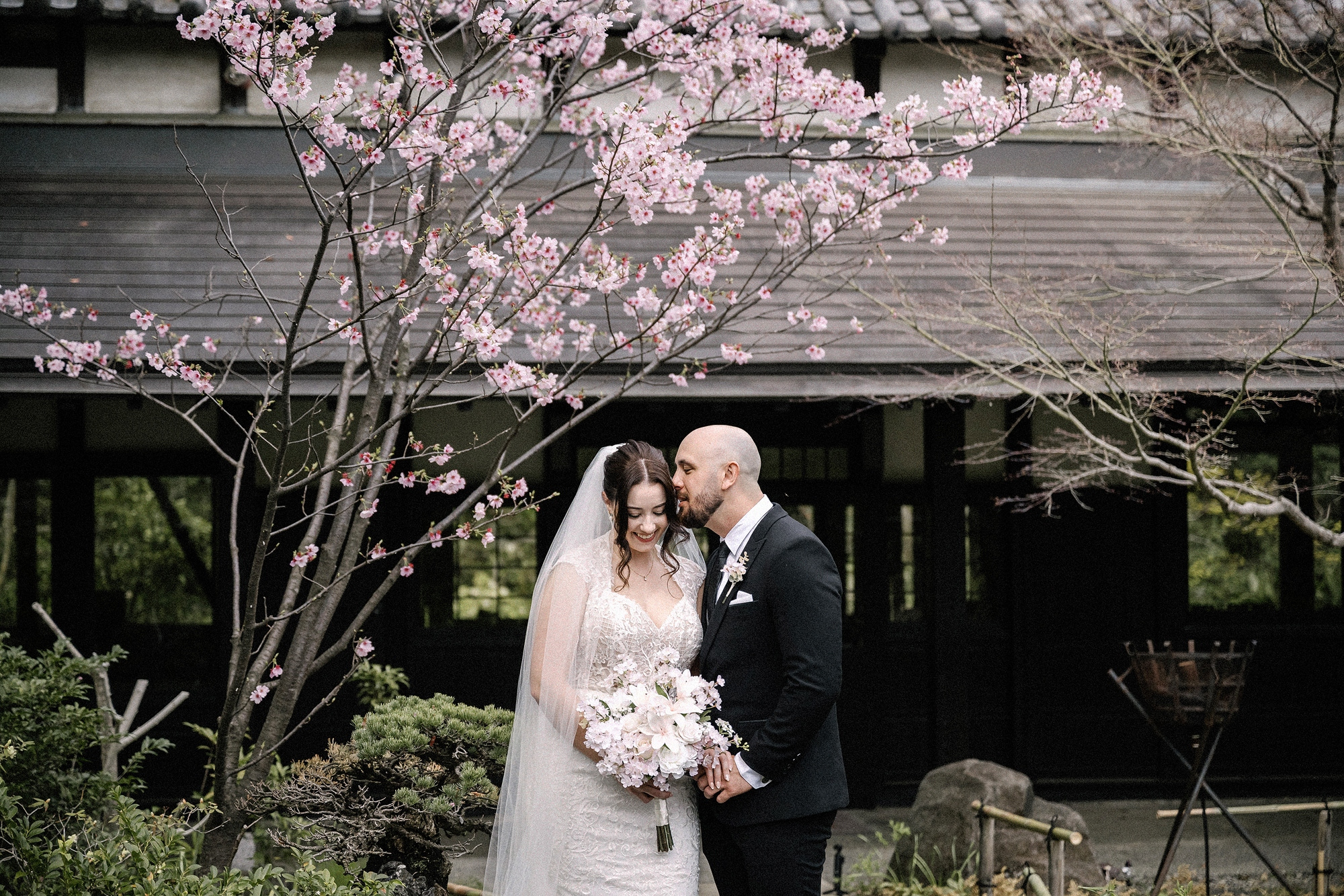 The groom kisses the bride's forehead under a pink cherry tree beside a dark timber hall, at their intimate spring wedding in Japan.