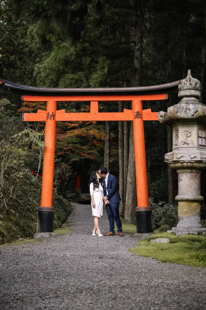 The couple kiss beneath a vermilion torii on a gravel path through tall cedars and stone lanterns, the day before their elopement in Japan.