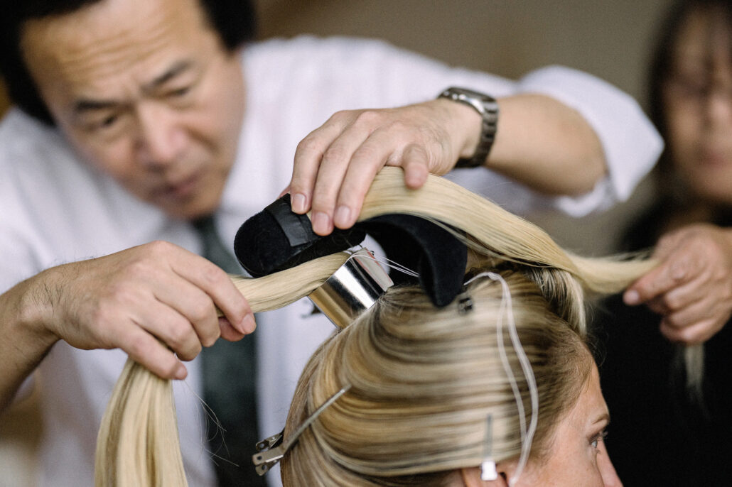 A nihongami master shapes a bride’s own blonde hair over a padded form while an assistant holds a section of her hair beside him