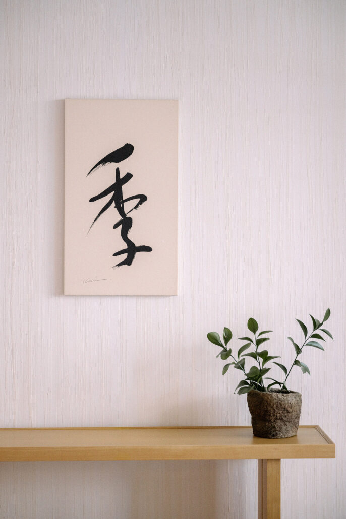 Brush calligraphy on a pale wall above a wooden console and a small potted plant, on the morning of a traditional Japanese wedding