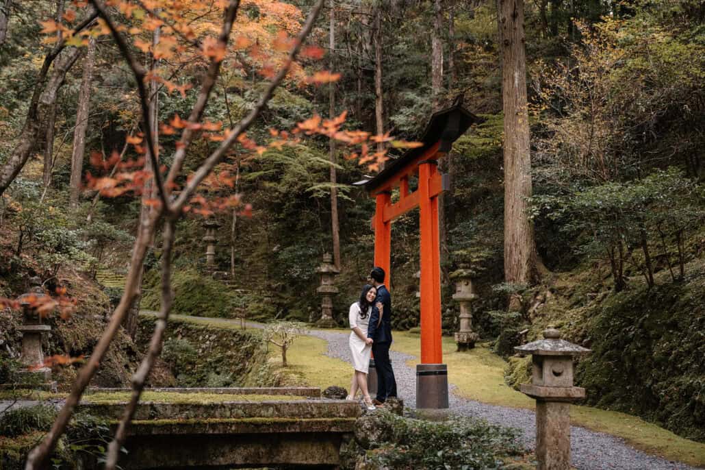The couple embrace under a vermilion torii among stone lanterns and moss, maple branches in red and orange in the foreground.