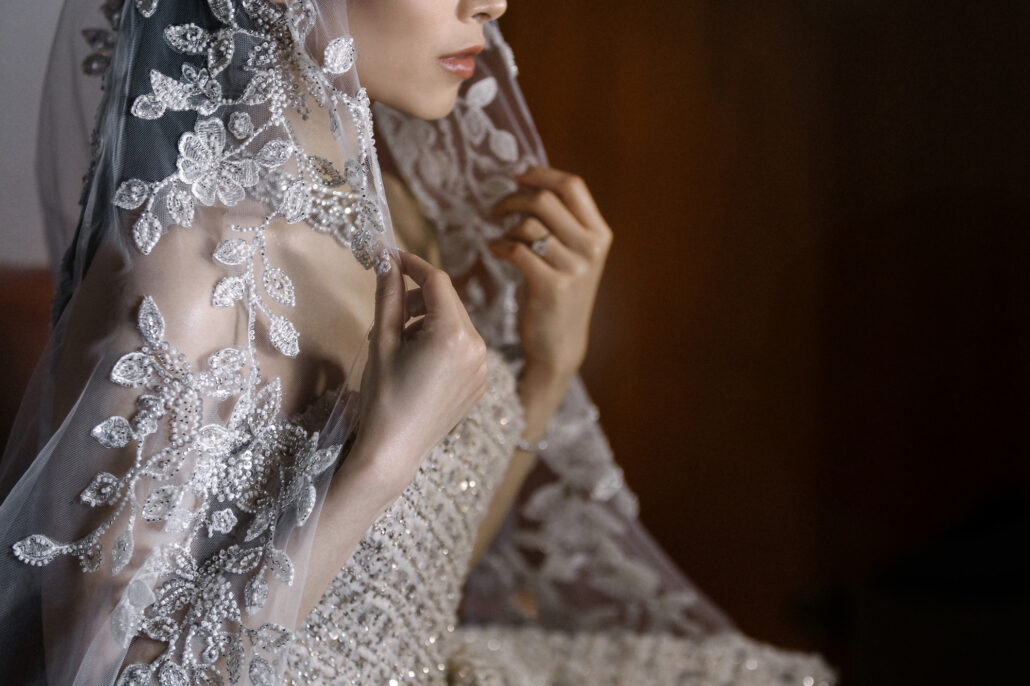 The bride's lace flower veil and beaded sleeve in warm light, her hand at her chin, a detail from the wedding day.