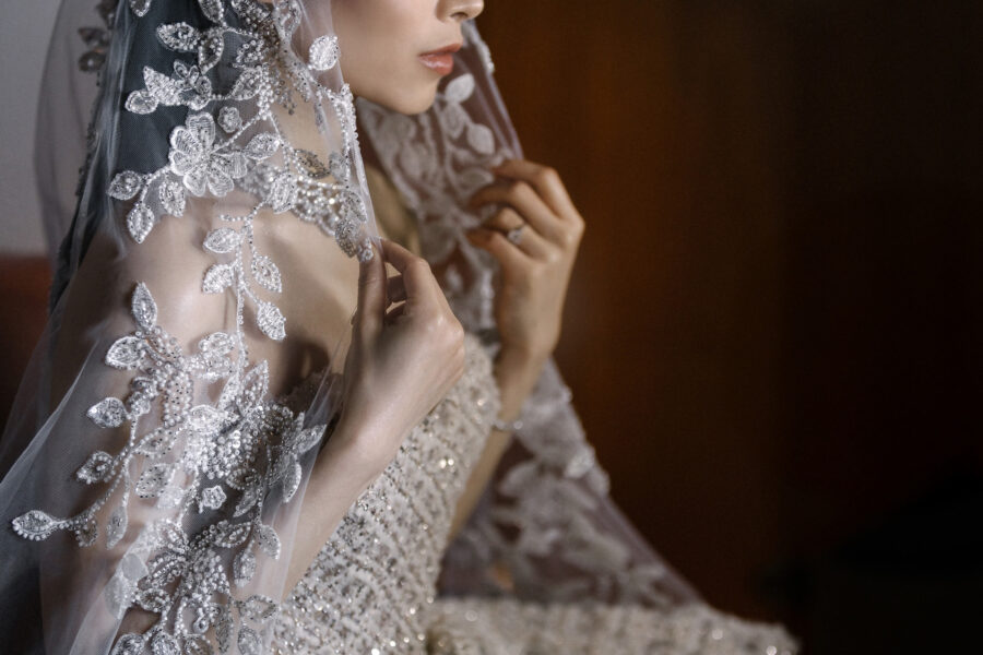 The bride's lace flower veil and beaded sleeve in warm light as she touches her lips, a detail from the wedding day.
