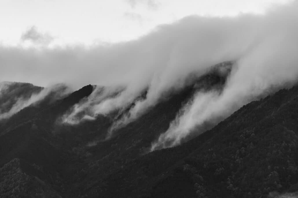 A black and white view of thick cloud spilling over the forested ridges above Lake Kawaguchiko on a wet late-autumn afternoon.