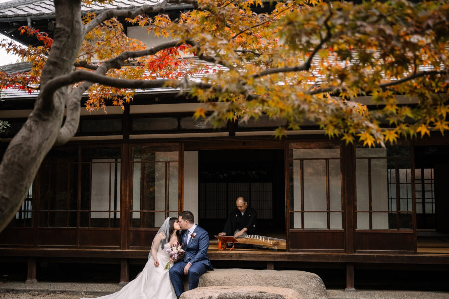 The couple sit together on a garden stone in autumn as a koto player plays on the open veranda of the wooden house.