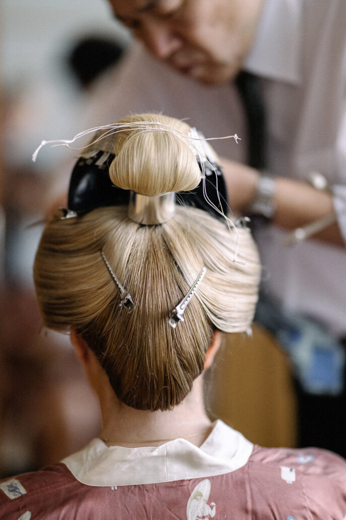 Back view of a bride’s nihongami taking shape, her blonde hair smoothed into a sleek curve at the nape and pinned around a topknot