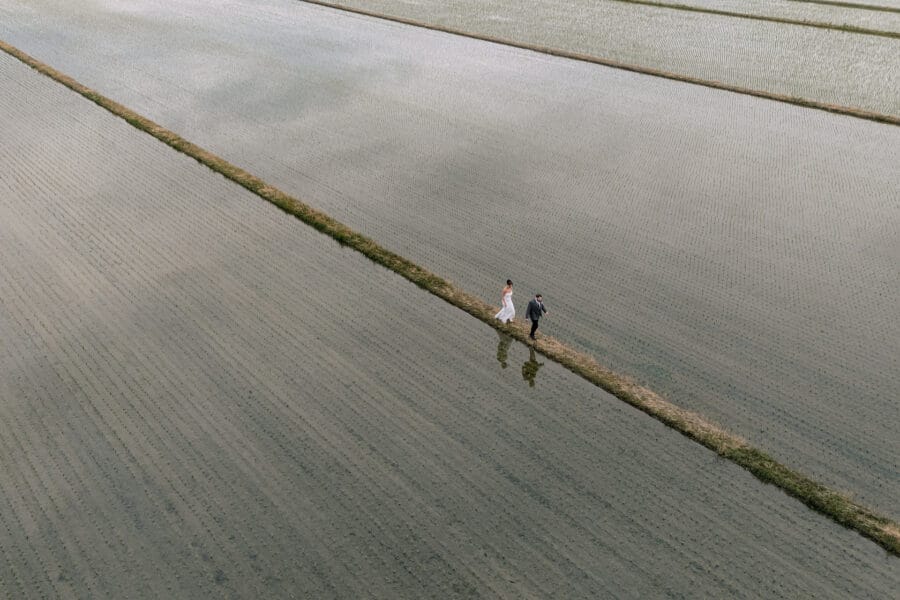 Seen from a drone, the newlyweds walk a narrow grass path between still, flooded rice paddies in Niigata, their reflections beside them.