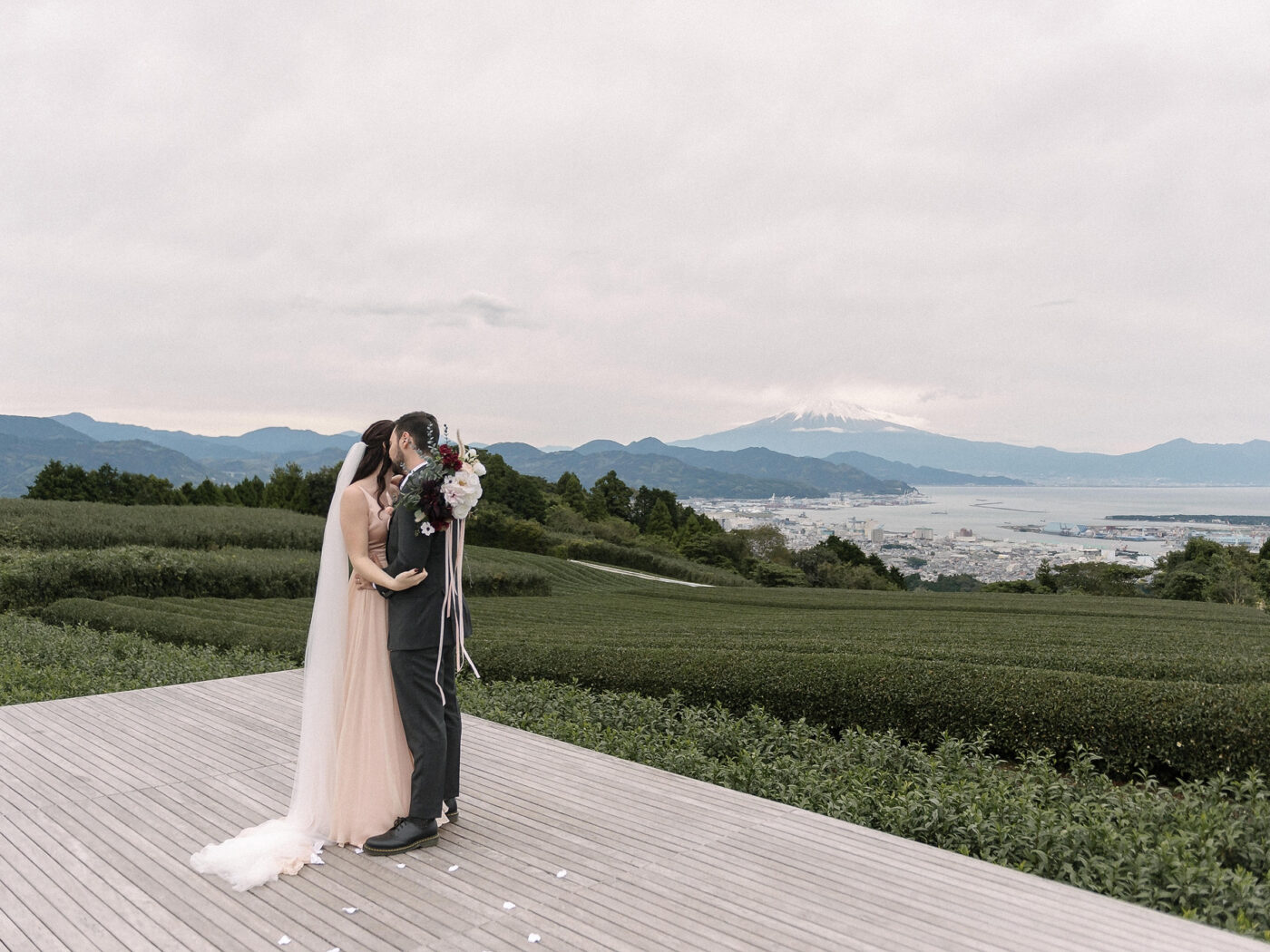 The couple kiss on the wooden tea deck at Nihondaira, rows of tea and the bay below, with Mt Fuji faint on the horizon.
