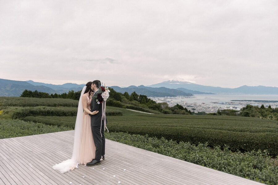 The couple kiss on the wooden tea deck at Nihondaira, rows of tea and the bay below, with Mt Fuji faint on the horizon.