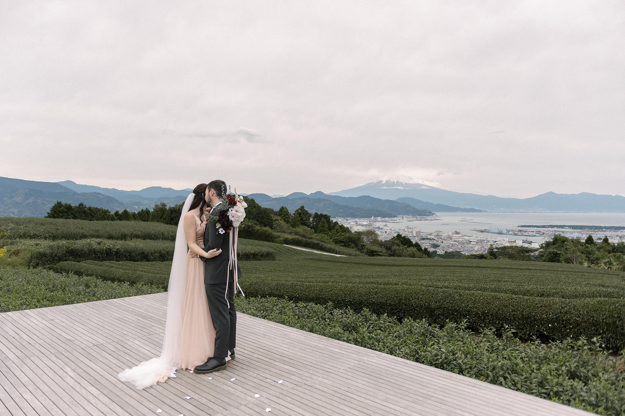 The couple kiss on the wooden tea deck at Nihondaira, rows of tea and the bay below, with Mt Fuji faint on the horizon.