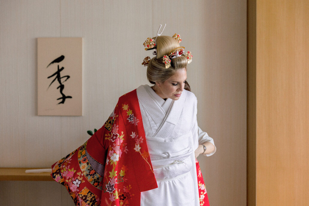 Bride in nihongami with flower kanzashi slipping a red iro-uchikake over her white kimono, beside a calligraphy panel in a hotel room