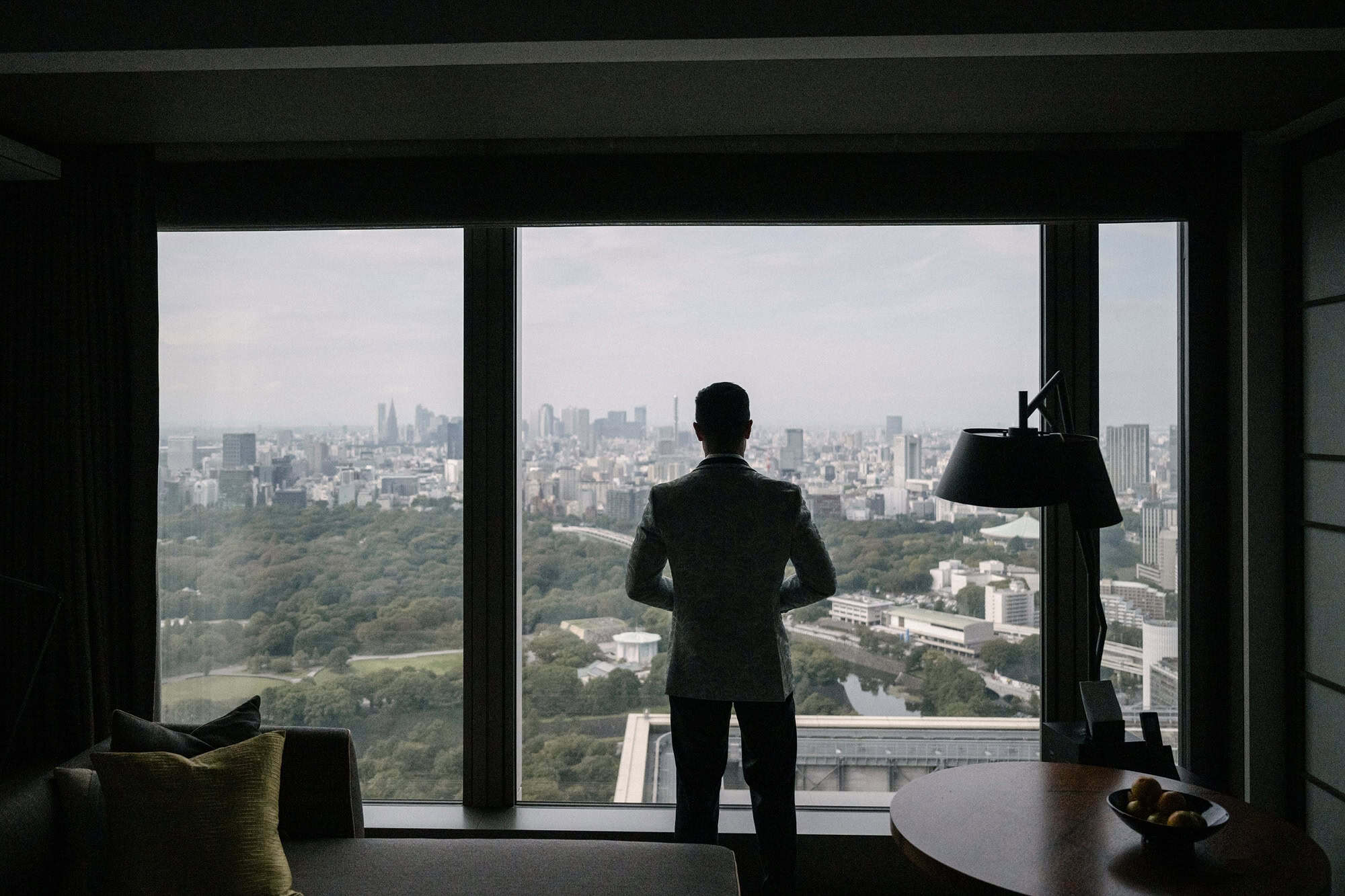 The groom stands alone at a tall window above a green park and the Tokyo skyline, getting ready for a hotel wedding in Tokyo.