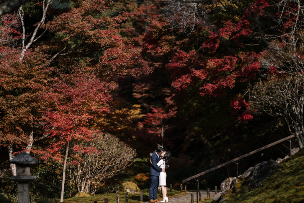 The couple kiss on a stone path beneath deep red maples at their Kyoto elopement, a stone lantern at the edge of the moss.